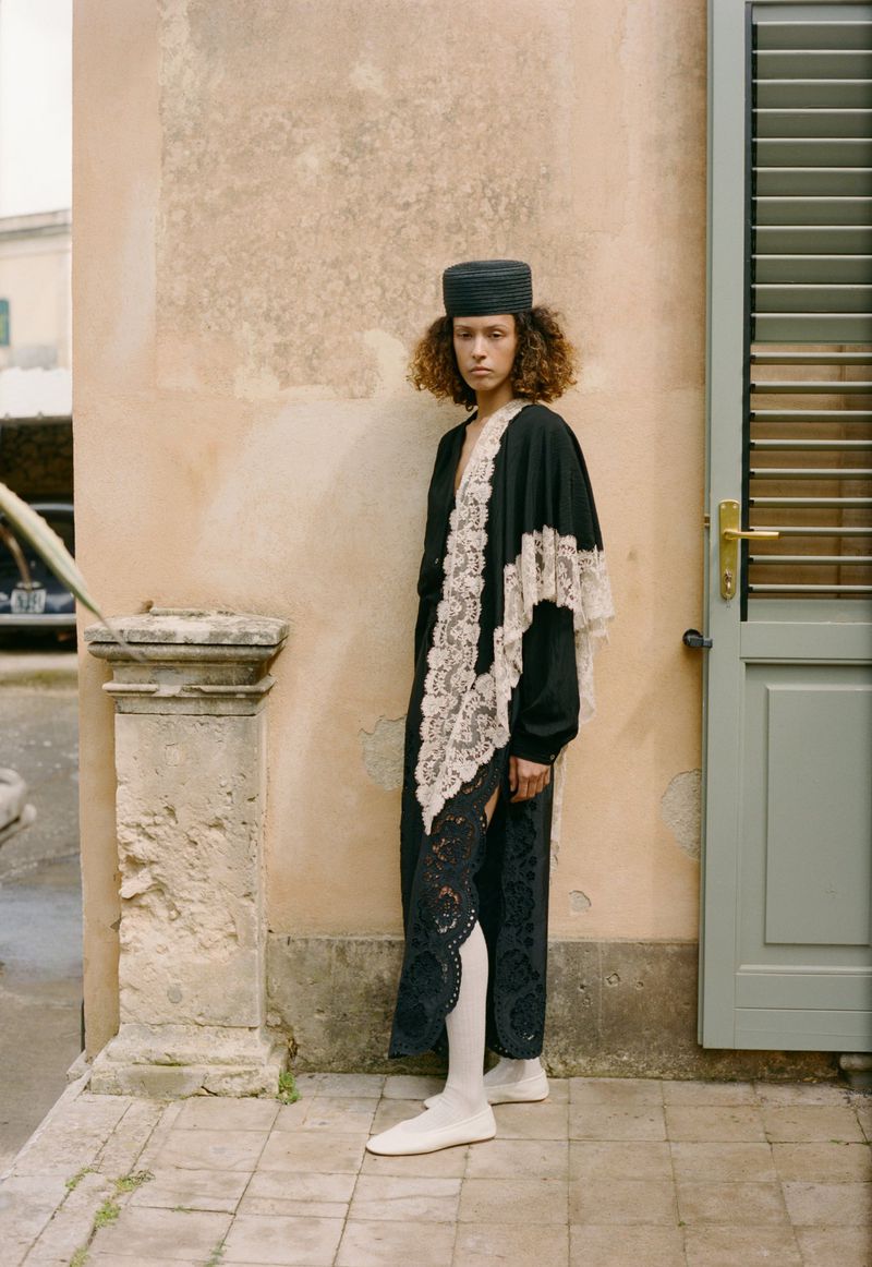 Model wearing a black lace-trimmed robe, black pillbox hat, white tights, and white slippers standing outside against a building wall.
