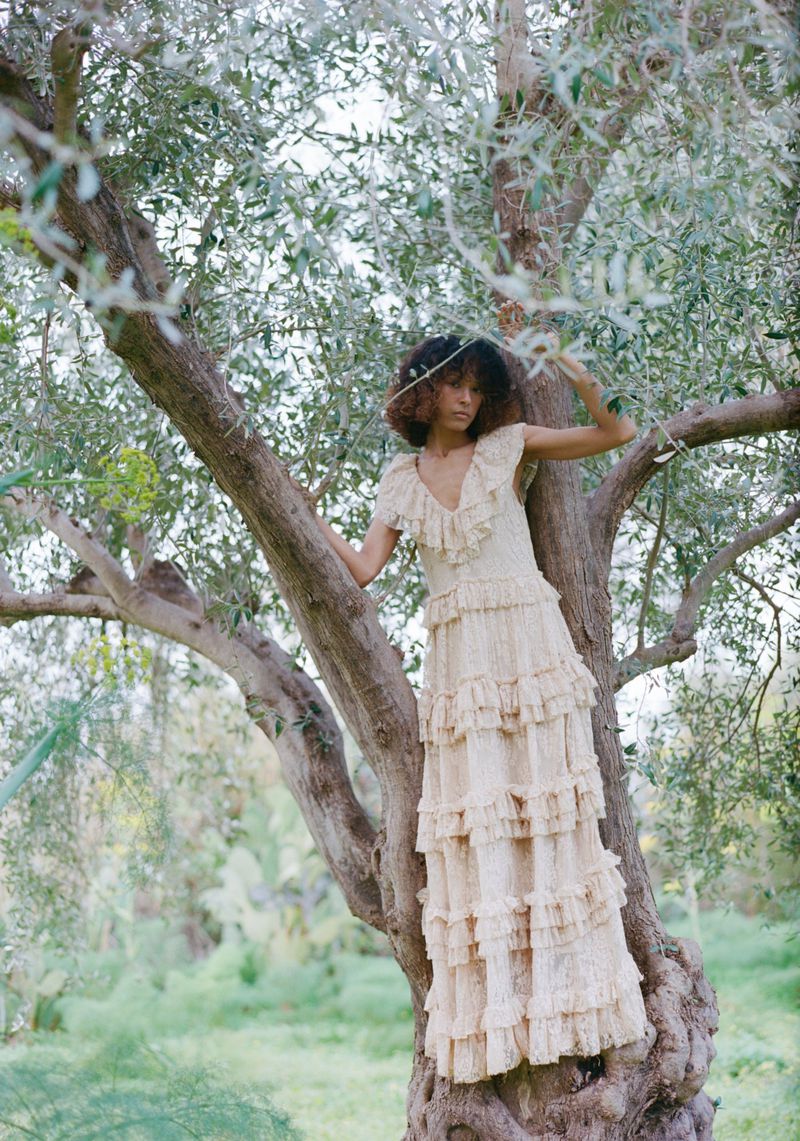 Fashion model wearing a tiered ruffle cream lace maxi dress while perched in a gnarled olive tree outdoors.