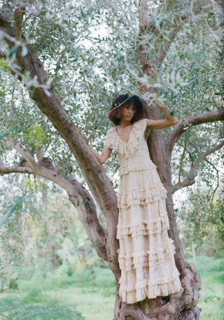 Fashion model wearing a tiered ruffle cream lace maxi dress while perched in a gnarled olive tree outdoors.