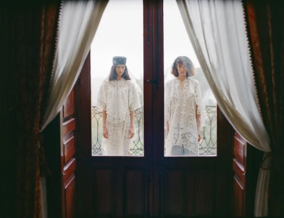 Two models wearing white lace garments posed on a balcony, viewed through wooden French doors and white drapes.