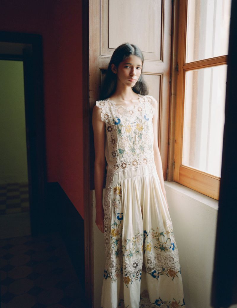 Model posing by a wooden window wearing an off-white embroidered lace dress in soft natural indoor light.