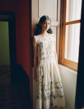 Model posing by a wooden window wearing an off-white embroidered lace dress in soft natural indoor light.