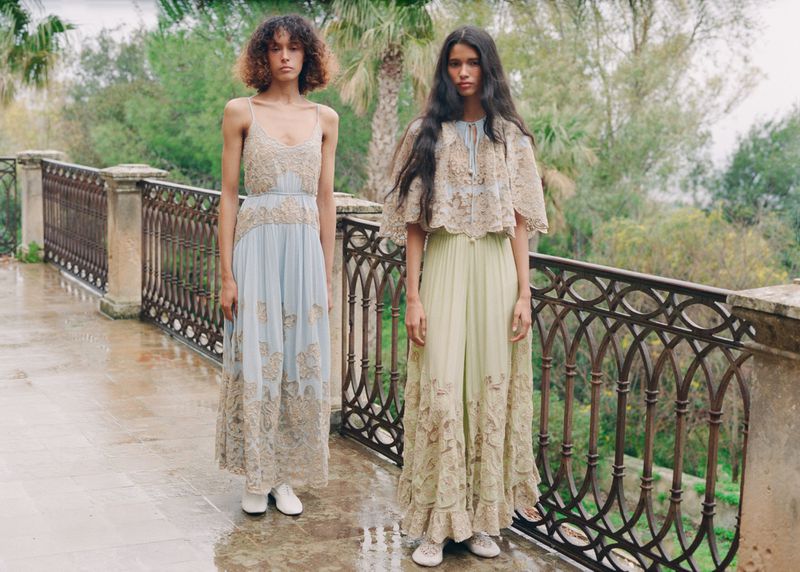 Two models standing on an outdoor stone terrace wearing lace-embellished maxi dresses with intricate embroidery by a wrought-iron railing.