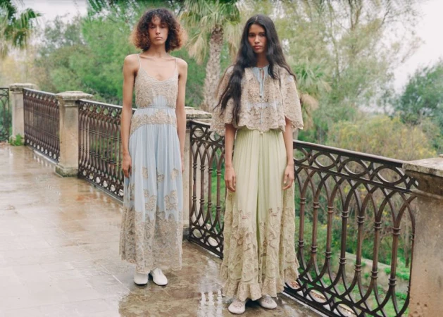 Two models standing on an outdoor stone terrace wearing lace-embellished maxi dresses with intricate embroidery by a wrought-iron railing.