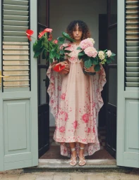 Model wearing a pink floral cape-sleeve maxi dress and holding potted anthurium and hydrangea plants in an open doorway.