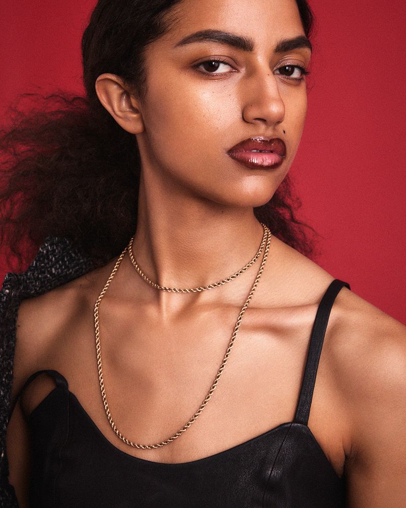 Close-up portrait of a woman wearing a double gold rope chain necklace, black strap top, and glossy dark lipstick against a red background.