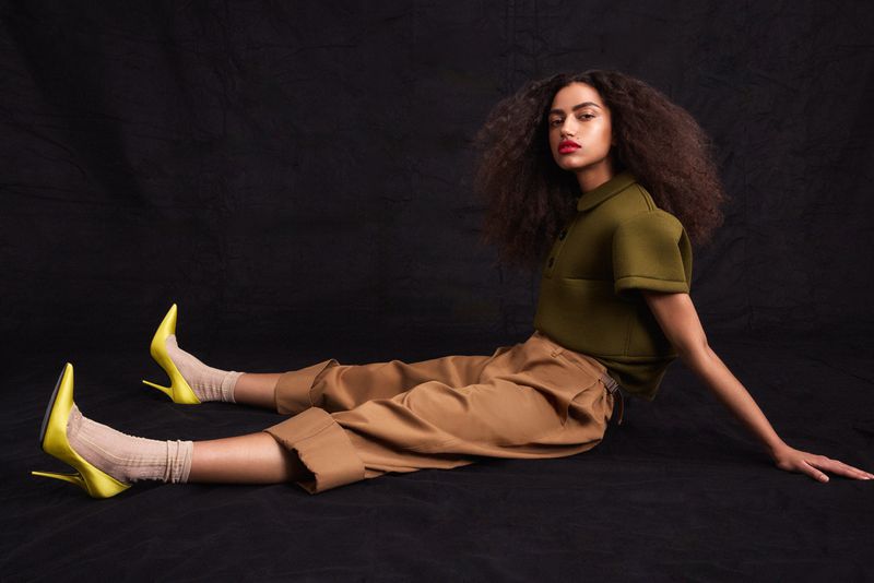 Woman with voluminous curly hair seated on a dark studio floor wearing an olive top, wide-leg camel trousers, and yellow stiletto heels.