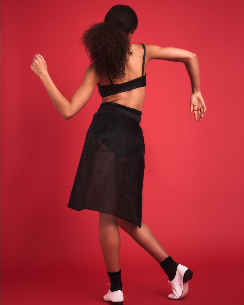 Model with dark curly hair in black top and skirt dancing against solid red background