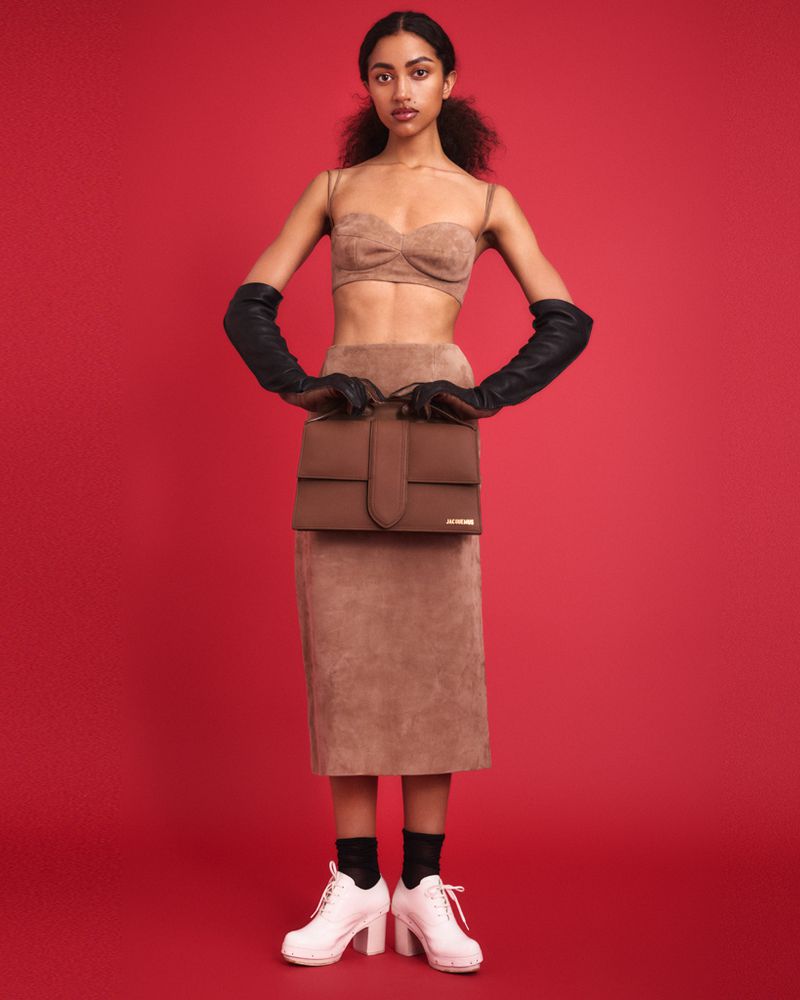 Model posing in a tan suede two-piece set, black opera gloves, and white platform shoes against a solid red studio background.