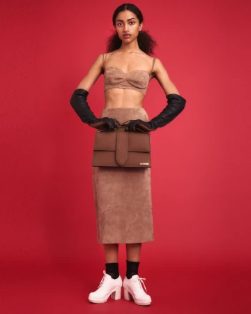 Model posing in a tan suede two-piece set, black opera gloves, and white platform shoes against a solid red studio background.