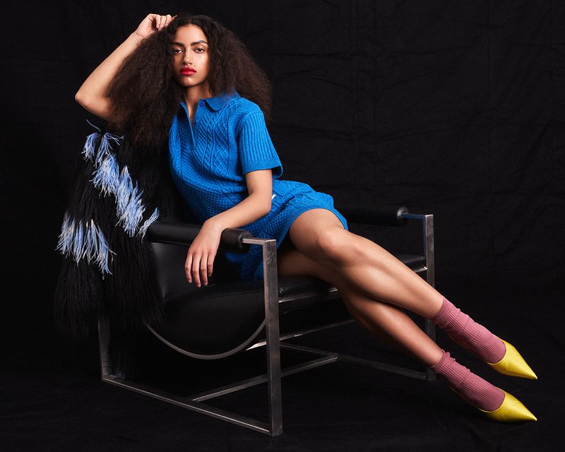 Model wearing a bright blue knit dress, pink socks, and yellow pointed heels reclined in a black leather armchair on a black background.