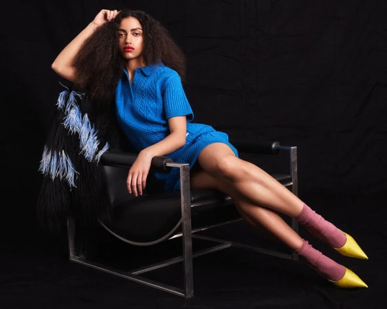 Model wearing a bright blue knit dress, pink socks, and yellow pointed heels reclined in a black leather armchair on a black background.