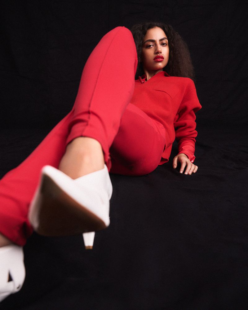 Model wearing a vibrant red collared shirt and red trousers with white pointed heels reclining against a black seamless backdrop.