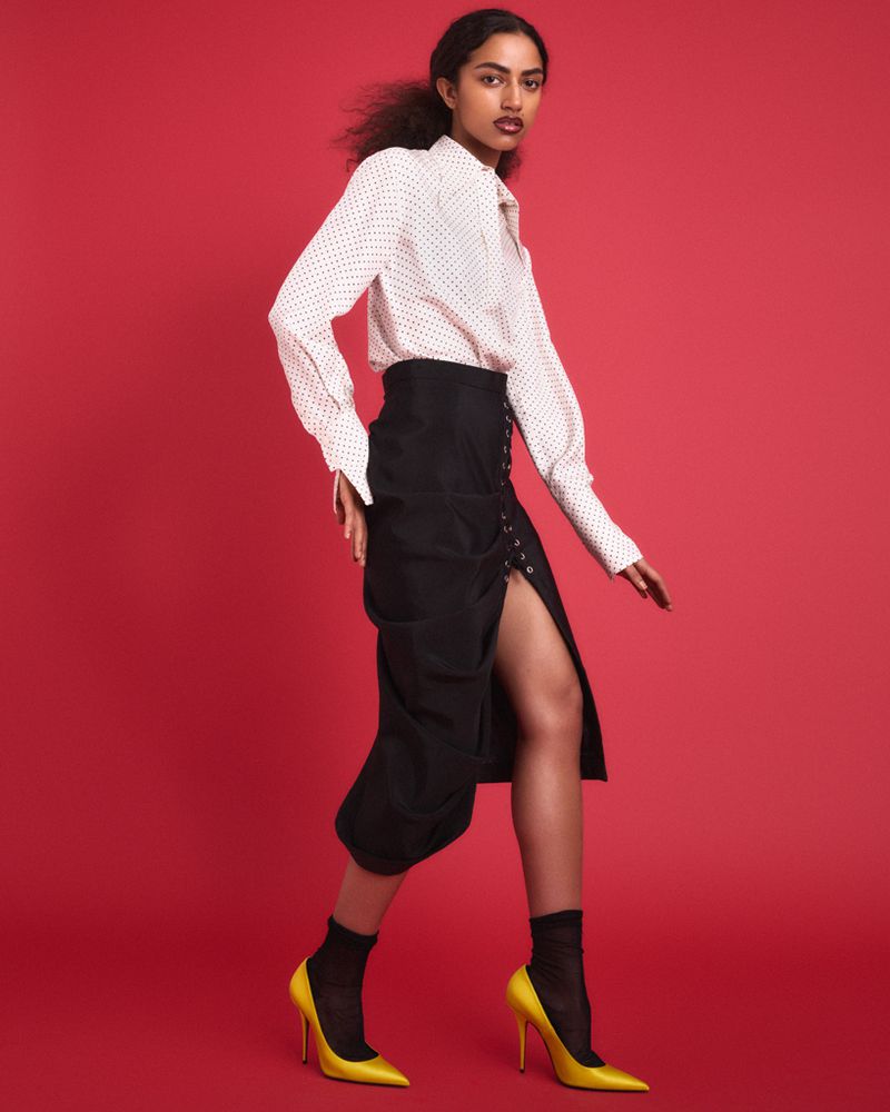 Model Isa Steadman Ruiz in a white patterned shirt, high-waisted black slit skirt, and yellow stiletto pumps against a red backdrop.
