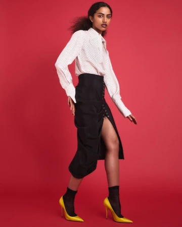 Model Isa Steadman Ruiz in a white patterned shirt, high-waisted black slit skirt, and yellow stiletto pumps against a red backdrop.