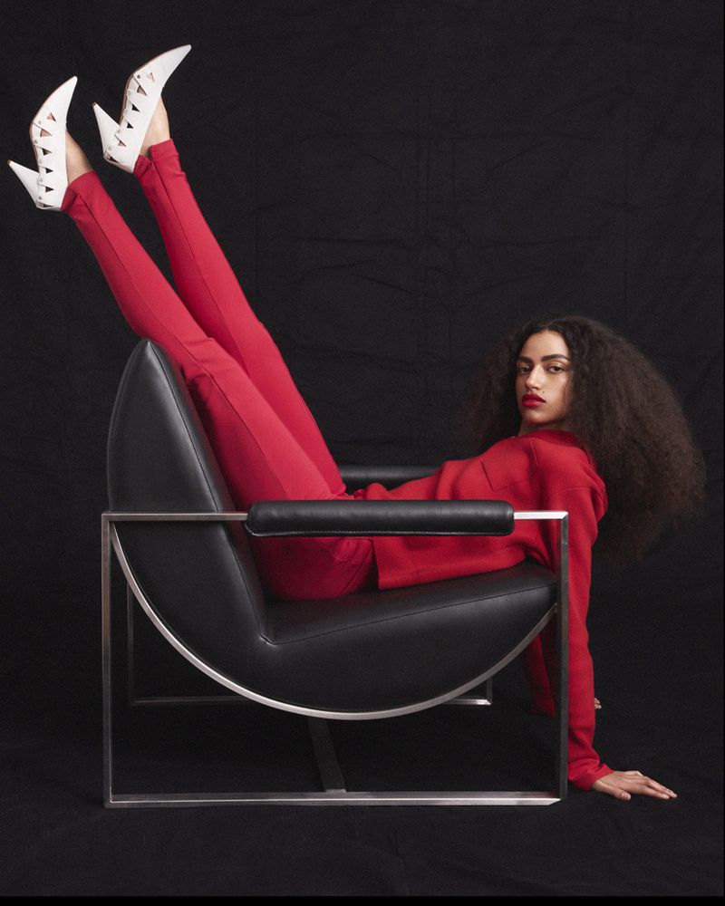 Woman in a red outfit and white cutout heels reclines upside down on a black leather chair against a dark backdrop.