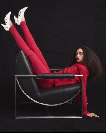 Woman in a red outfit and white cutout heels reclines upside down on a black leather chair against a dark backdrop.