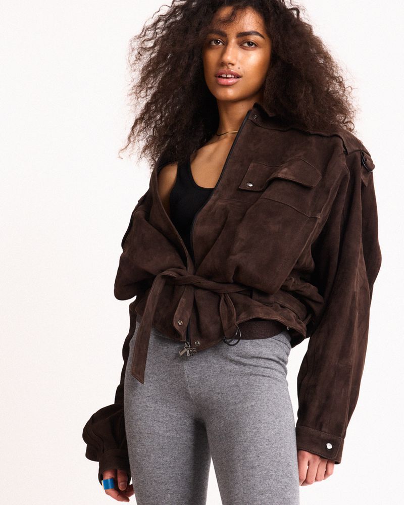 Fashion model with voluminous curly hair posing in an oversized brown suede belted jacket and grey leggings against a white backdrop.