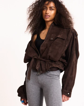 Fashion model with voluminous curly hair posing in an oversized brown suede belted jacket and grey leggings against a white backdrop.