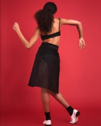 Model with dark curly hair in black top and skirt dancing against solid red background