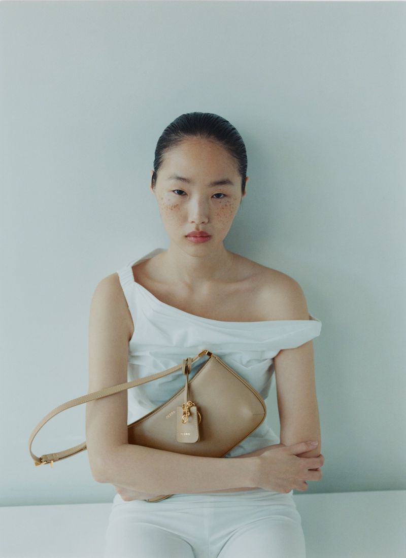 East Asian woman with freckles wearing a white off-shoulder top and holding a beige leather shoulder bag against a plain wall.