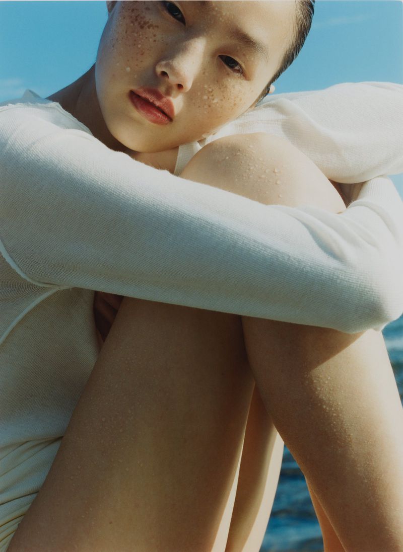 Close-up portrait of an East Asian woman with freckles wearing a white long-sleeve knit top with a blue ocean backdrop.