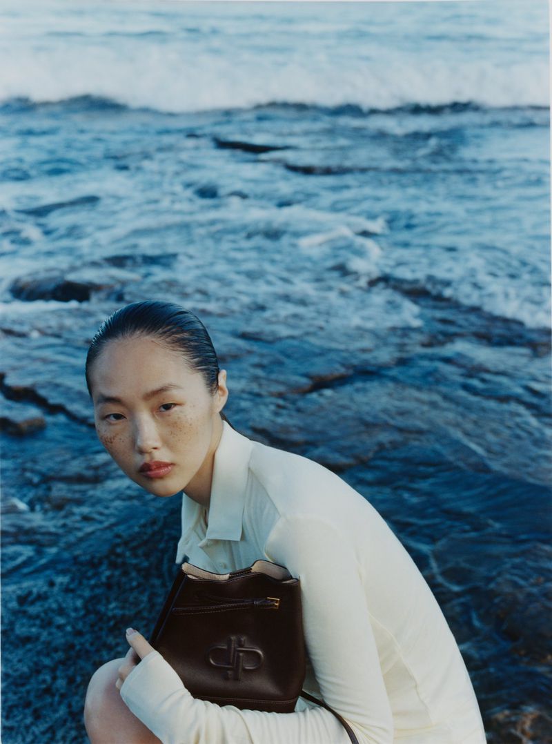 Model Peng Chang crouching near ocean waves holding a dark brown leather bucket bag for the PEDRO The Splash campaign.