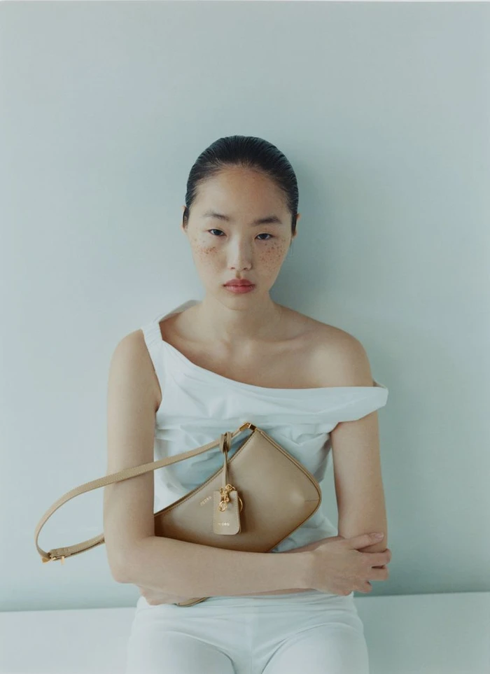 East Asian woman with freckles wearing a white off-shoulder top and holding a beige leather shoulder bag against a plain wall.