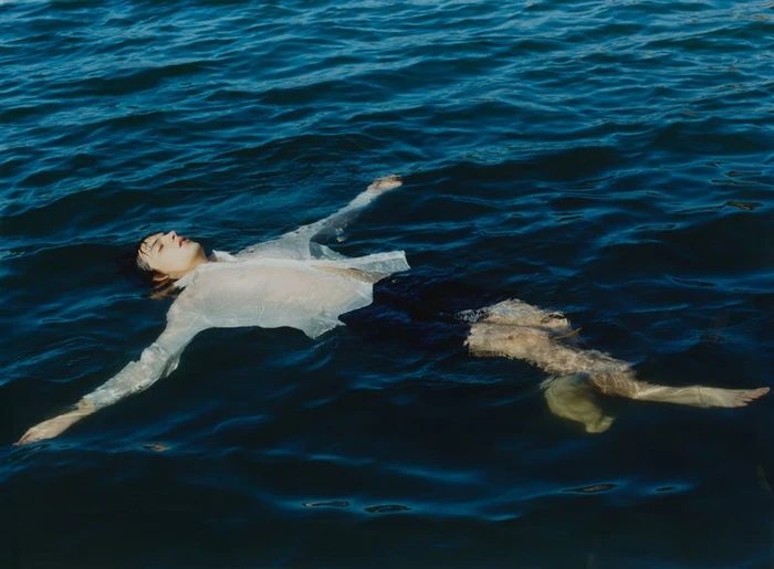 A man wearing a sheer wet white button-down shirt floats on his back in deep blue open water.
