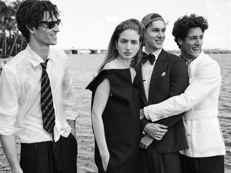 Four models posing outdoors by the water wearing monochrome formal tailoring and dresses in a black and white editorial portrait.