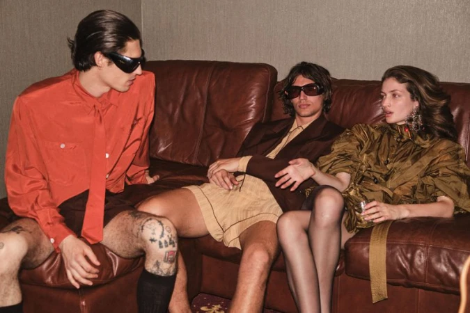 Three models seated on a brown leather sofa in an indoor lounge setting wearing tailored attire and sunglasses.