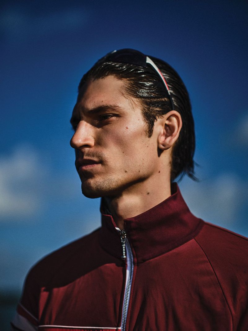 Close-up portrait of a male model wearing a burgundy zip-up jacket against a clear blue sky background.