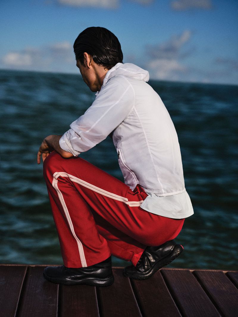 Model crouching on a wooden pier wearing a white hooded jacket and red track pants against an ocean background