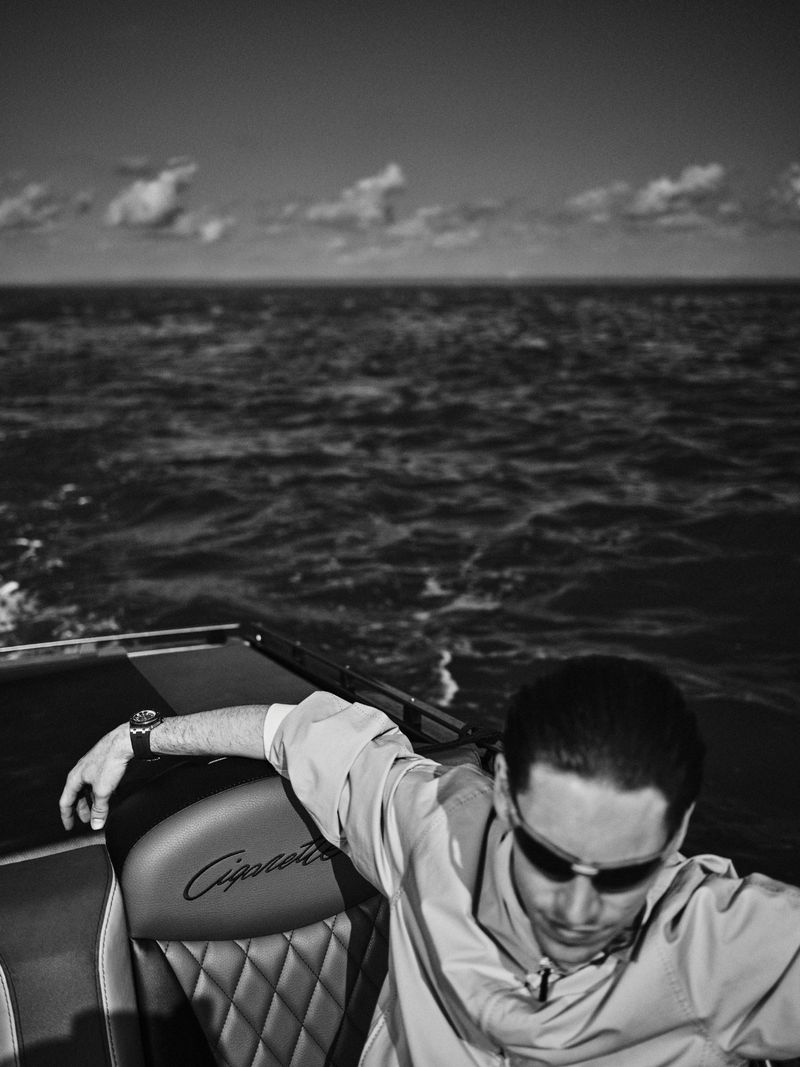 Justin Eric Martin wearing dark sunglasses and a light jacket seated in a speedboat on the ocean in black and white.