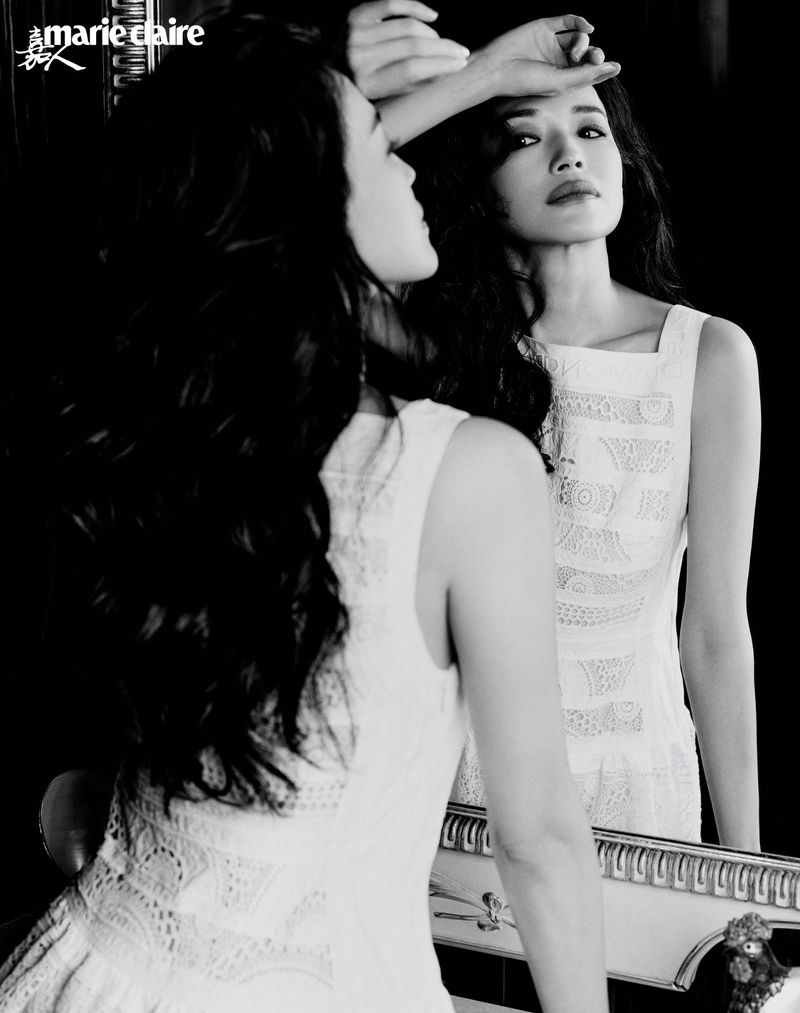 Actor Shu Qi wearing a white sleeveless lace dress, leaning forward and reflected in an ornate mirror in a monochrome studio setting.