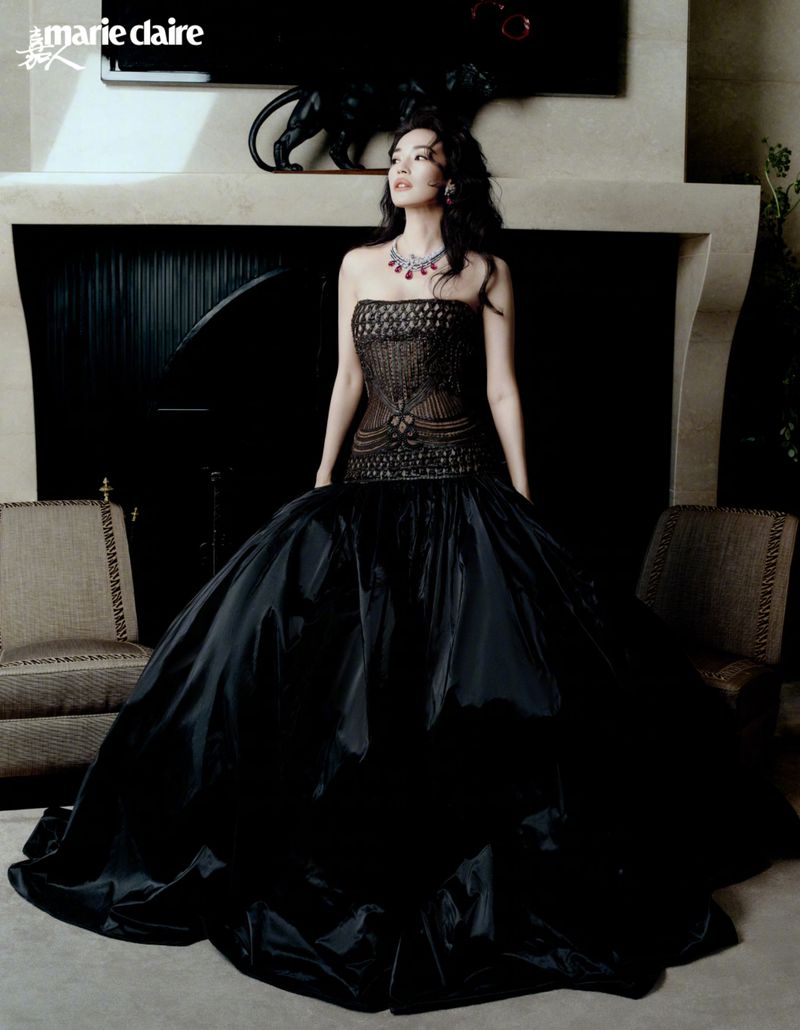 Shu Qi posing in a voluminous black strapless ball gown and statement gemstone necklace in front of a stone fireplace.
