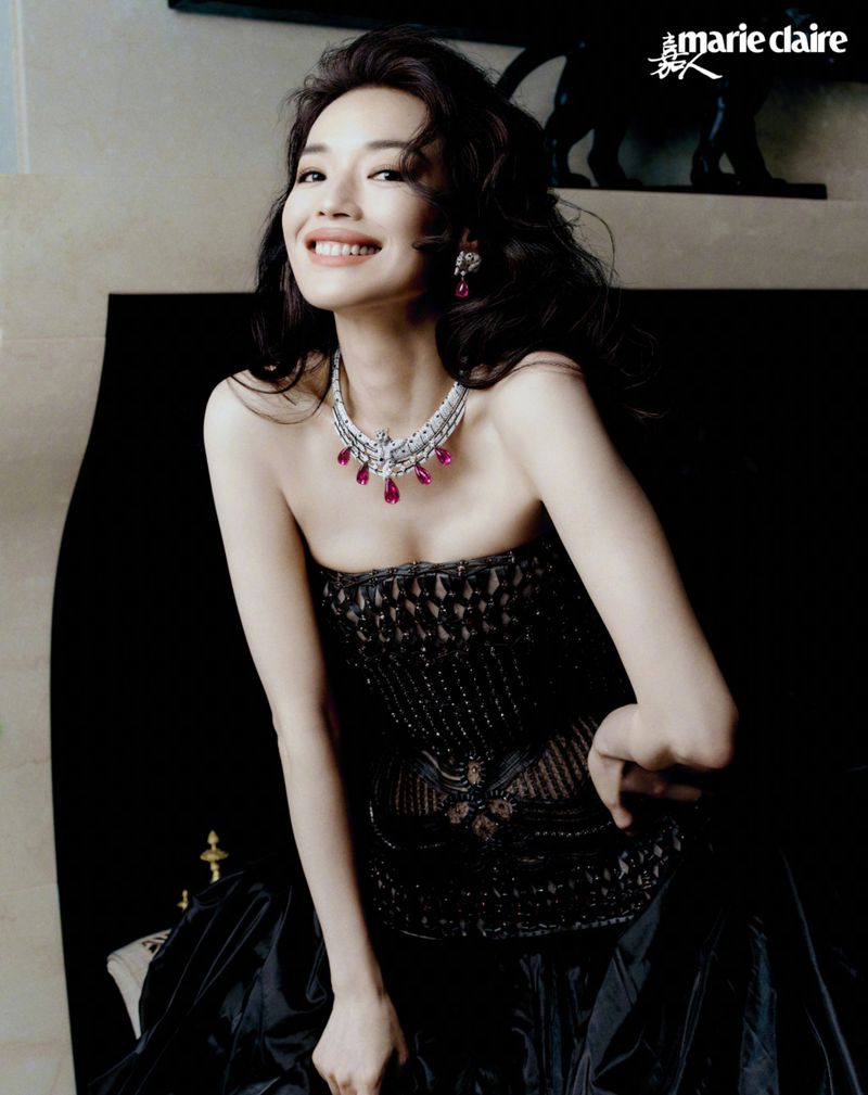 Shu Qi smiling in a beaded black strapless gown and diamond necklace with ruby drop pendants in a dimly lit interior setting.