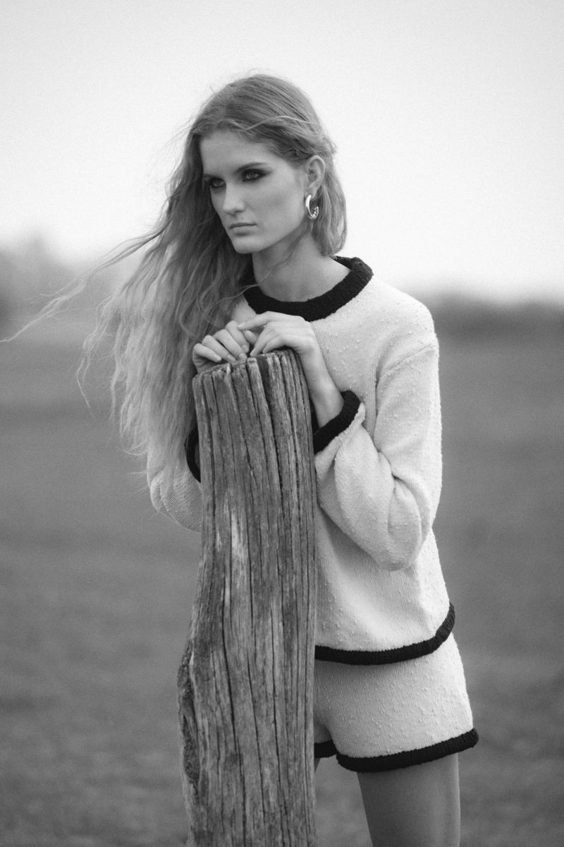Monochrome portrait of a woman in a knit sweater and matching shorts leaning against a weathered wooden post in an open field.