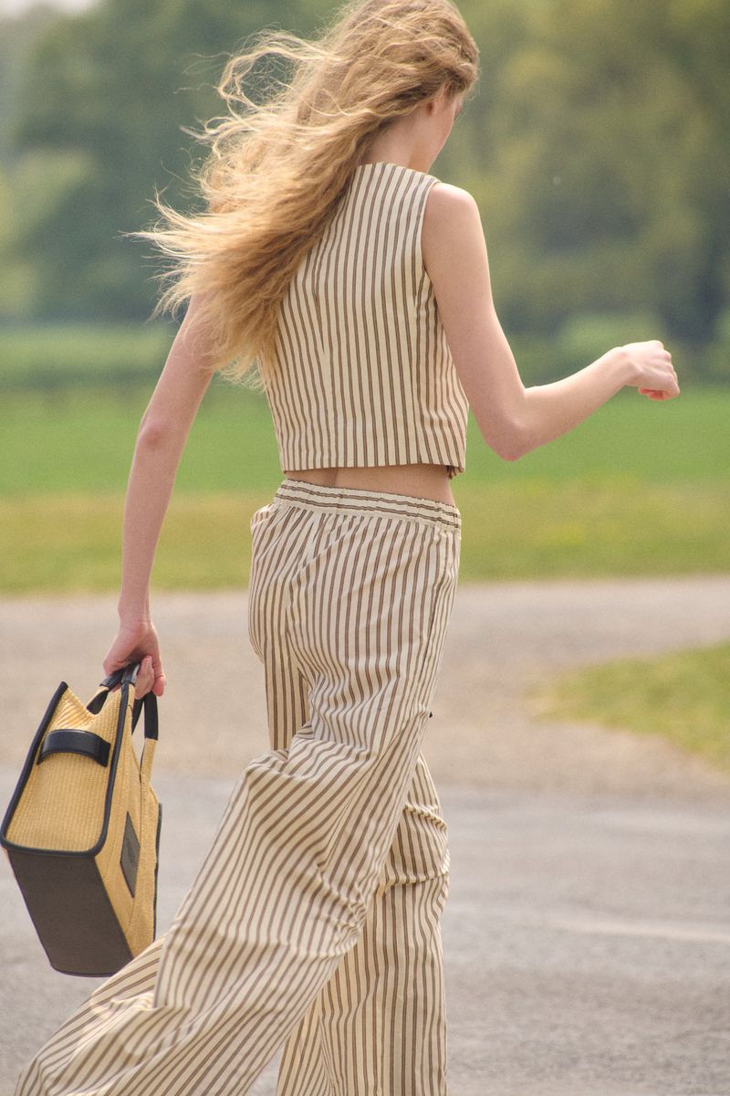 Back view of a woman with windblown blonde hair walking in a striped crop top and wide-leg trousers while carrying a tote bag.