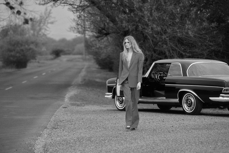 Model wearing a tailored trouser suit walking on a gravel roadside beside a classic black coupe in black and white.