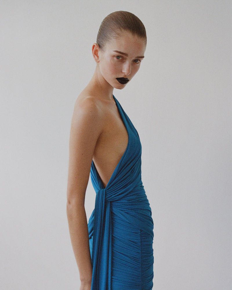 A fair-skinned woman wearing a blue dress with dark lipstick poses in a studio, captured in a medium shot against a neutral background.