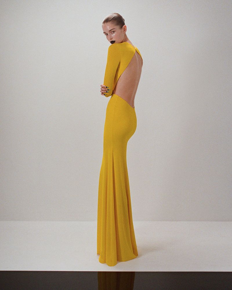 Elegant woman in a yellow gown with an open back posing in a studio with a minimalist white background and a black reflective floor.