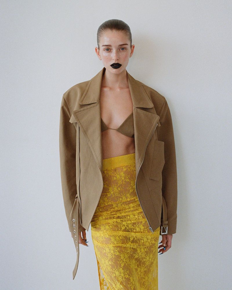 Woman in a tan jacket and yellow skirt poses against a white wall, showcasing modern fashion with a minimalist studio backdrop.