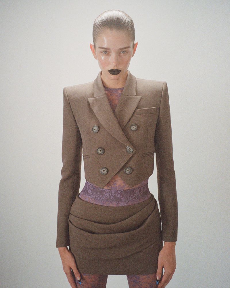 Woman poses in a cropped brown blazer and matching mini skirt against a white backdrop, showcasing fashion and style with striking makeup.
