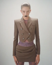 Stella Hanan wearing a cropped taupe double-breasted blazer and matching draped mini skirt against a neutral studio backdrop.
