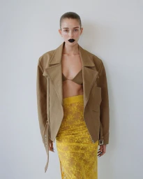 Stella Hanan wearing an oversized tan jacket and yellow lace skirt against a plain white studio background.