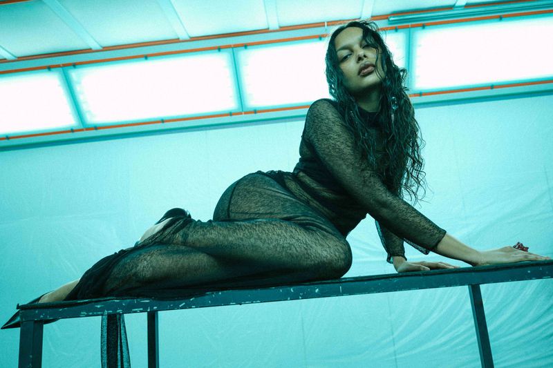 Model in sheer black long-sleeve dress reclining on a dark metal platform under cyan studio lighting.