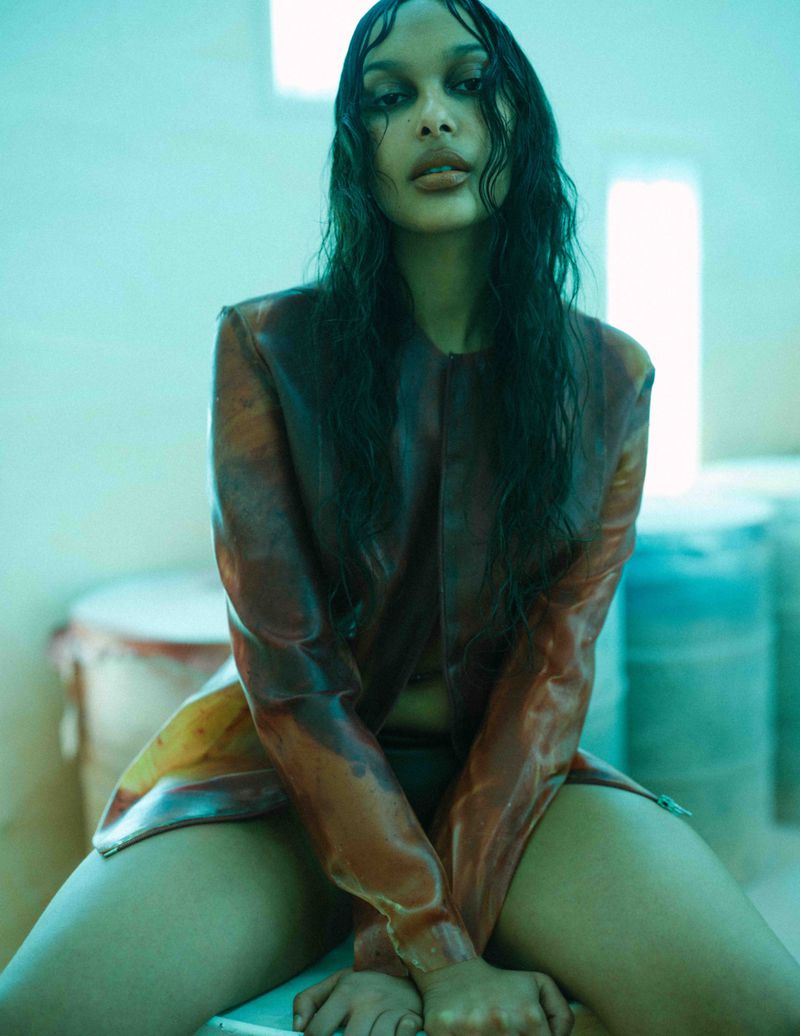 Fashion portrait of a woman with wet-look dark hair wearing a glossy brownish jacket, seated in a cool-toned ambient setting.