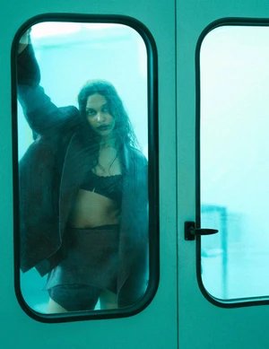 Woman in an oversized dark jacket and bralette looking through a door window in cool cyan lighting.