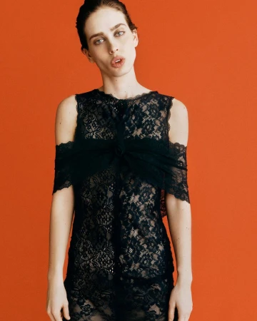 Model Sean Levy in a sheer black lace cold-shoulder dress posing against a solid vibrant orange seamless studio background.